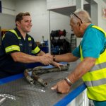 Two male workers in safety vests shaking hands over a workbench in a hydraulic franchise workshop in Baltimore, MD.