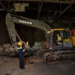 A construction worker in high-visibility gear speaks with an operator inside a Volvo excavator in a dimly lit industrial services building.