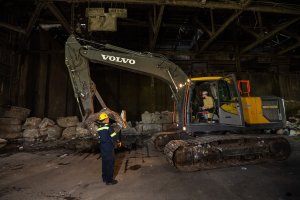 A construction worker in high-visibility gear speaks with an operator inside a Volvo excavator in a dimly lit industrial services building.