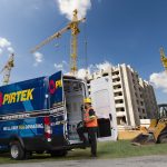 Construction worker in reflective vest standing by a Pirtek franchise opportunities service van at a site with cranes and under-construction building.