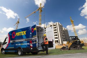 Construction worker in reflective vest standing by a Pirtek franchise opportunities service van at a site with cranes and under-construction building.