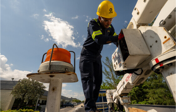 A worker in safety gear, including a yellow helmet, stands on a hydraulic utility vehicle and repairs or inspects electrical equipment under a clear sky.